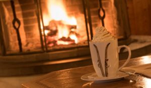 Eine Tasse heiße Schokolade mit Sahne auf einem Holztisch vor einem Kaminfeuer.